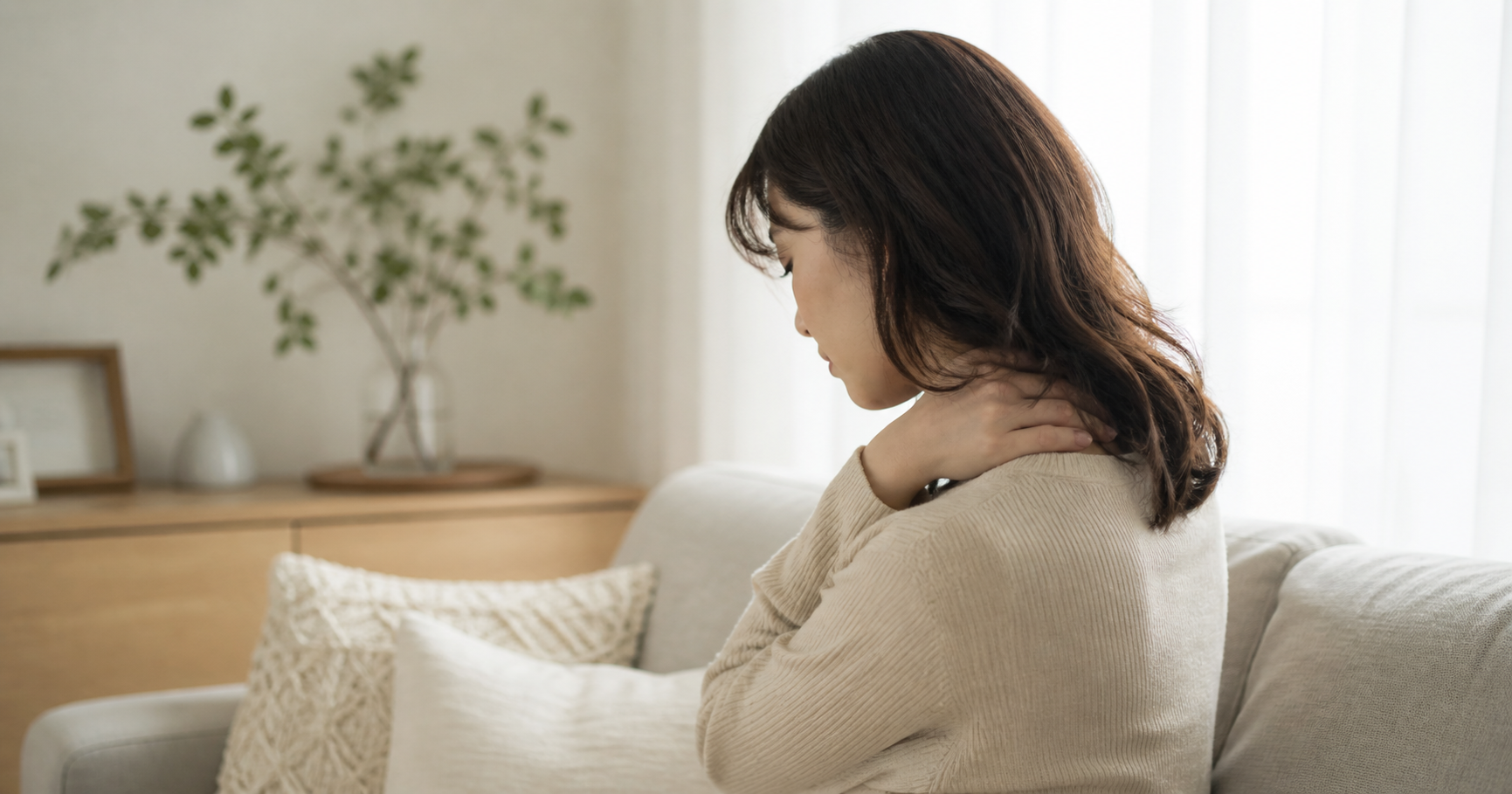 白とベージュを基調にした明るい室内で、女性がソファに浅く座り、首から肩に手を添えながらうつむいている様子